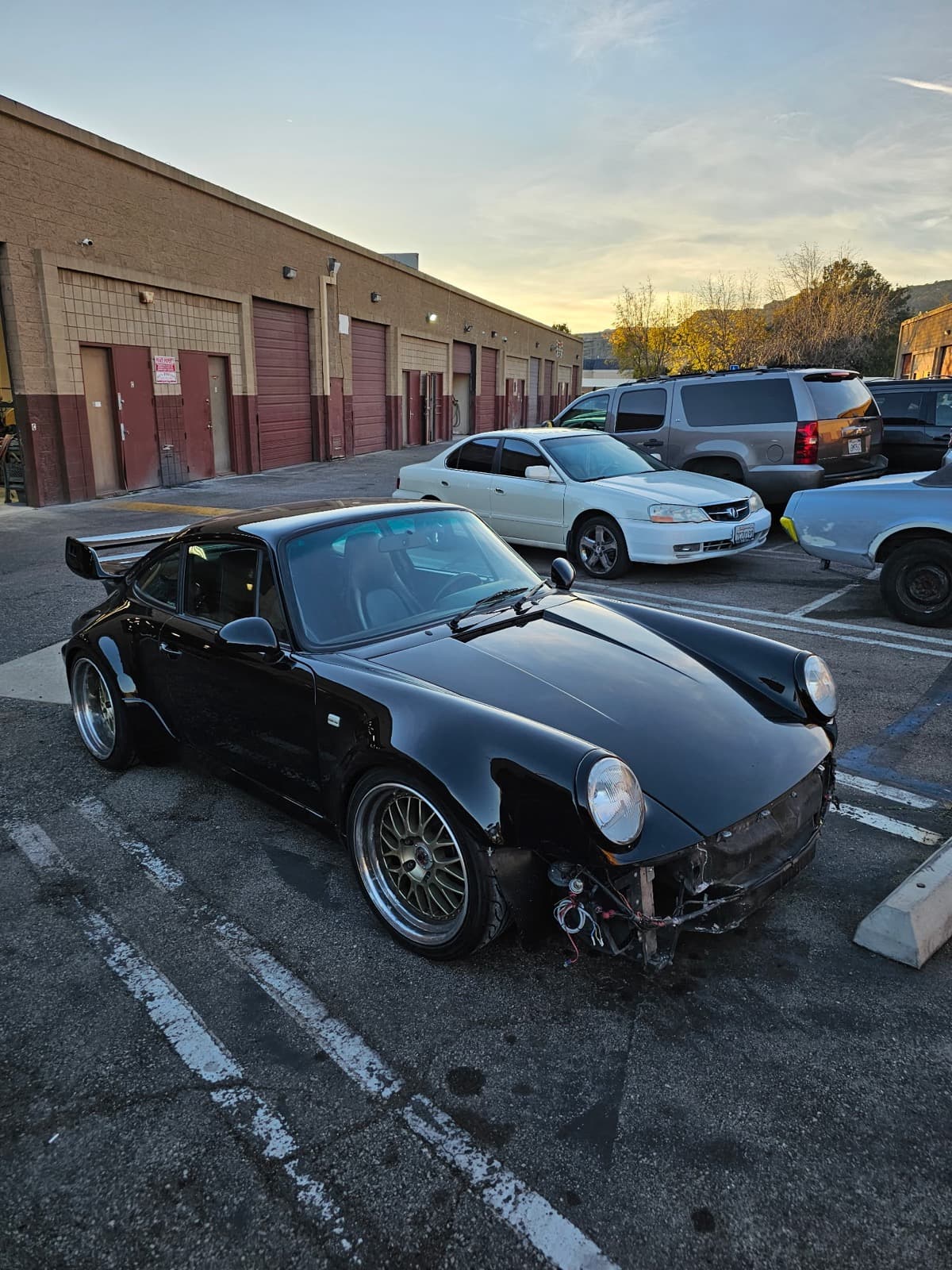 A black classic air-cooled Porsche 911 with a whale-tail spoiler and widebody fenders, its front bumper removed for collision repair, parked in the shop's lot