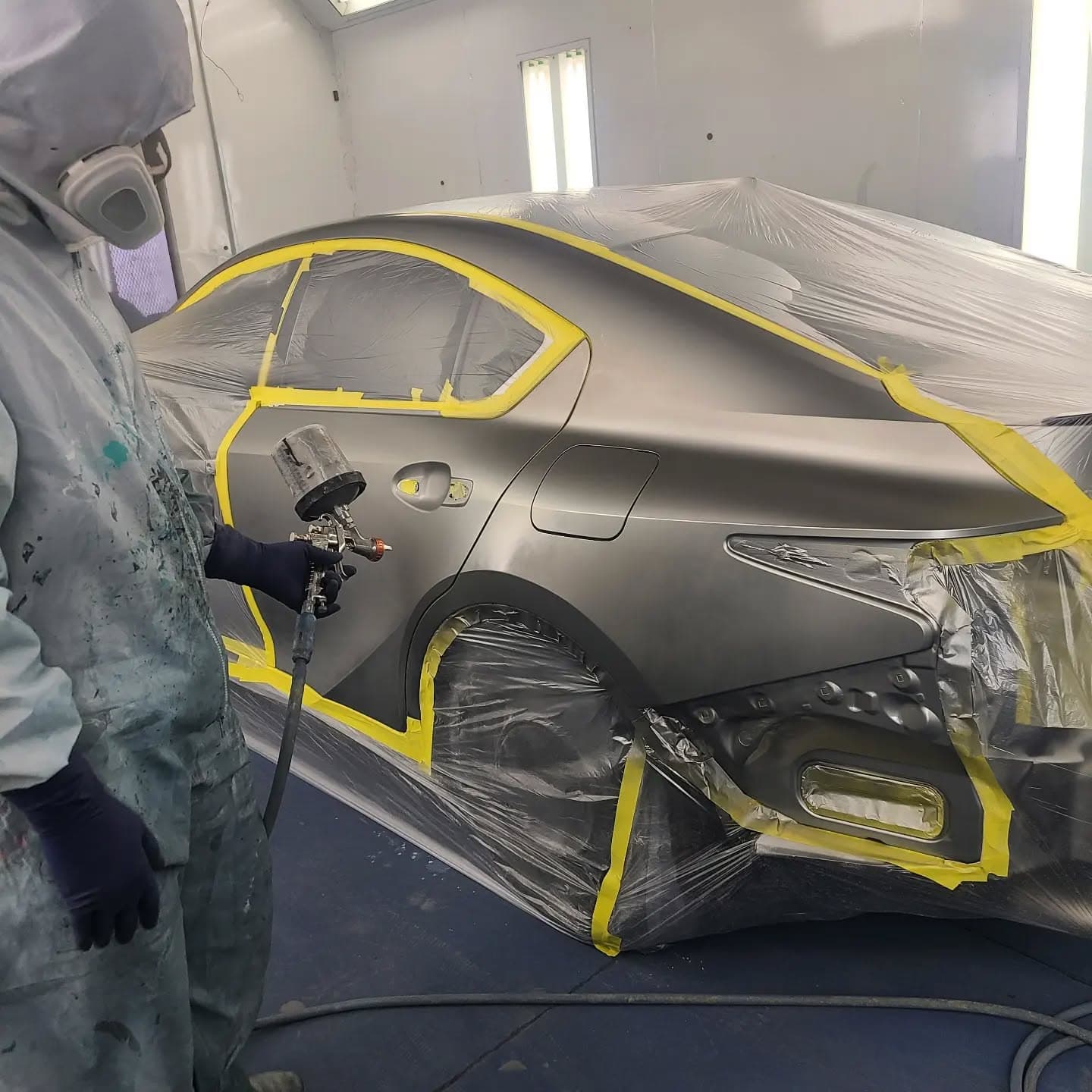 A painter in a protective suit and respirator spraying fresh color onto a masked grey car inside the shop's spray booth