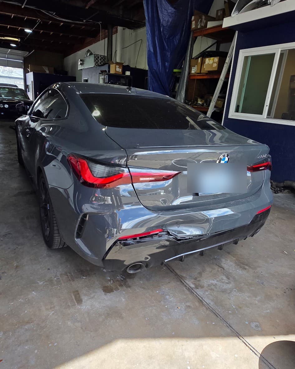 A gunmetal-grey BMW 430i seen from the rear inside the shop bay, its rear bumper cover and diffuser in for repair