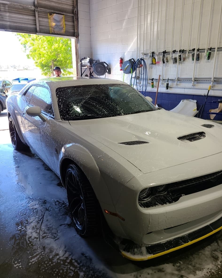 A grey Dodge Challenger inside the shop's wash bay, front three-quarter view, its windshield and body covered in wash foam before pickup